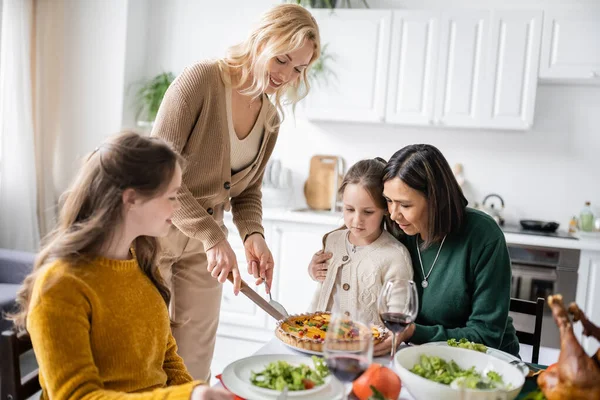 Donna sorridente taglio torta di ringraziamento vicino multietnico madre e figlia a casa — Foto stock