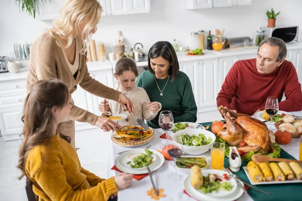 Famille multiculturelle avec des enfants regardant la femme couper tarte thanksgiving pendant le dîner à la maison — Photo de stock