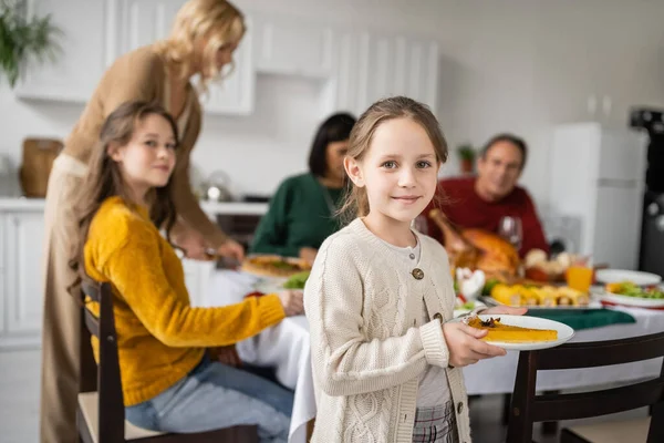 Fille tenant une tarte savoureuse près de la famille floue célébrant le dîner de Thanksgiving à la maison — Photo de stock