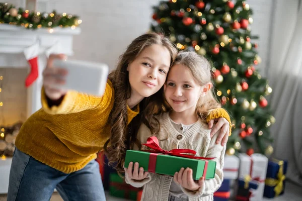 Улыбающийся подросток делает селфи с сестрой, держащей подарок на Рождество дома — стоковое фото