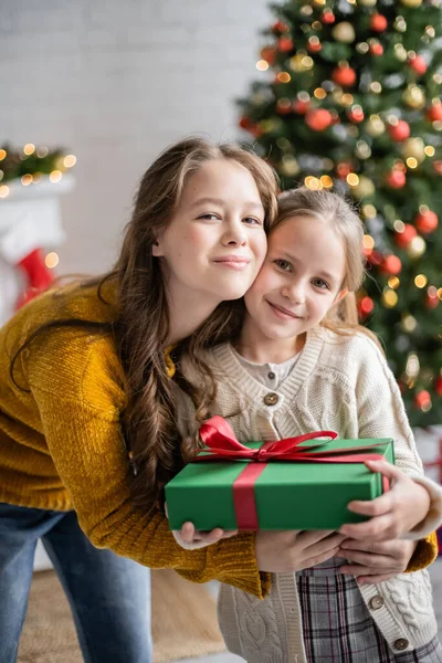 Sorridente adolescente abbracciare sorella con presente durante il Natale a casa — Foto stock