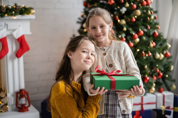 Glückliche Schwestern mit Geschenkschachtel, die in die Kamera neben dem geschmückten Weihnachtsbaum auf verschwommenem Hintergrund schauen — Stockfoto