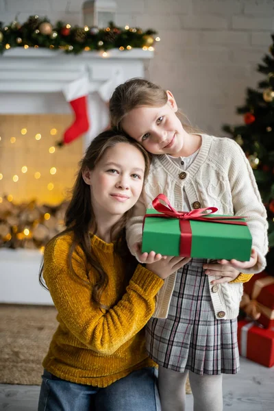 Lächelnde Schwestern mit Geschenkbox und Blick in die Kamera im Wohnzimmer mit Weihnachtsdekoration — Stockfoto