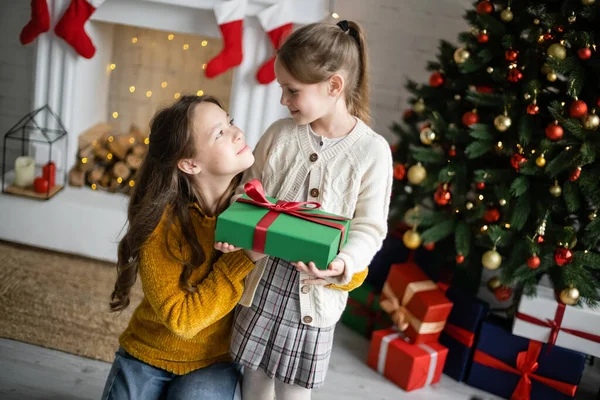 Irmãs felizes segurando caixa de presente e olhando uns para os outros perto de lareira borrada e árvore de natal na sala de estar — Fotografia de Stock