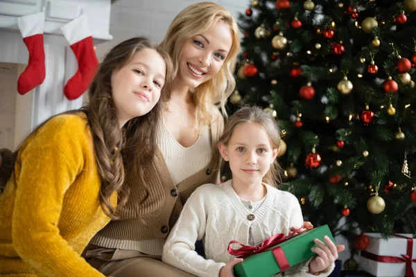 Chicas felices con madre rubia mirando a la cámara cerca del árbol de Navidad decorado sobre fondo borroso - foto de stock