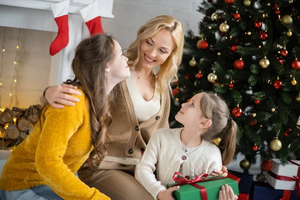 Sonriente madre abrazando hijas cerca decorado árbol de Navidad y chimenea sobre fondo borroso - foto de stock