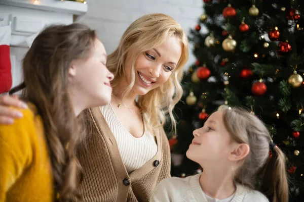 Mujer rubia feliz mirando a las hijas cerca del árbol de Navidad decorado sobre fondo borroso - foto de stock