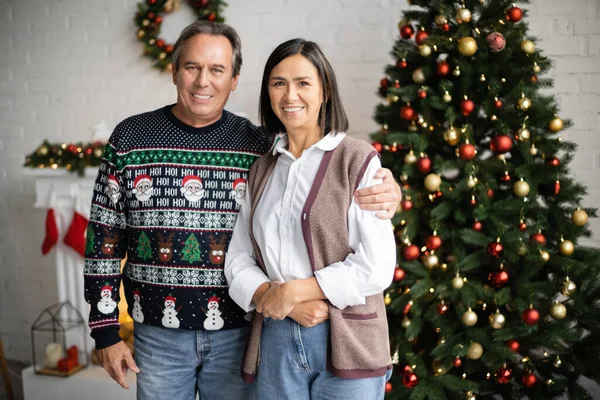 Homem sorridente em suéter com padrão festivo abraçando esposa multirracial perto de árvore de natal borrada — Fotografia de Stock