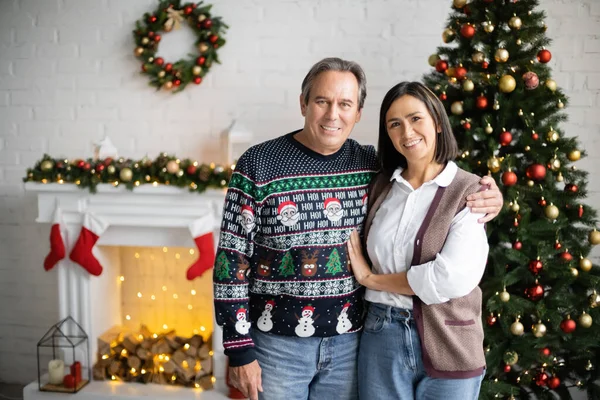 Heureux couple multiethnique souriant à la caméra dans le salon avec cheminée décorée et arbre de Noël — Photo de stock