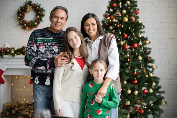 Smiling sisters looking at camera near happy interracial grandparents and christmas tree in living room — Stock Photo