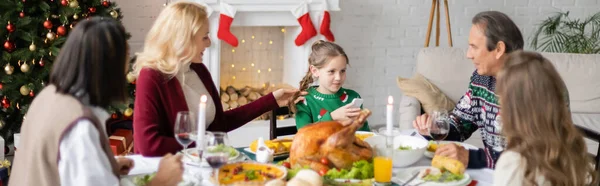 Дівчина тримає мобільний телефон біля матері і міжрасових бабусь і дідусів під час святкування Різдва, банер — стокове фото