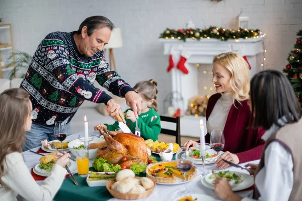 Пожилой человек режет жареную индейку рядом с межрасовой семьей на рождественский ужин дома — стоковое фото