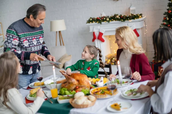 Улыбающаяся девушка смотрит на дедушку с ножом во время рождественского ужина с межрасовой семьей — стоковое фото