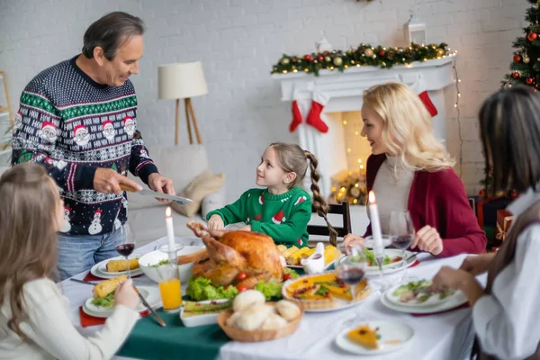 Smiling girl looking at grandfather with knife while having christmas dinner with interracial family — Stock Photo