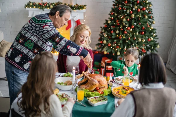 Hombre mayor cortando delicioso pavo asado durante la cena familiar cerca del árbol de Navidad - foto de stock