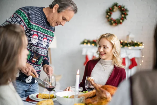 Uomo anziano taglio mais alla griglia e sorridente vicino alla famiglia avendo cena di Natale — Foto stock