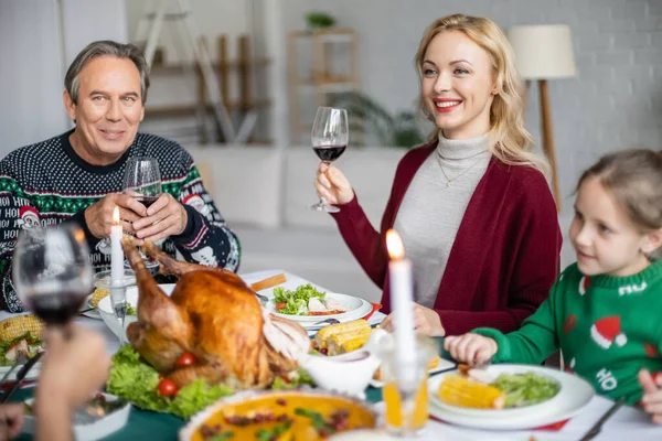 Cheerful woman toasting with glass of red wine near family and delicious thanksgiving dinner — Stock Photo