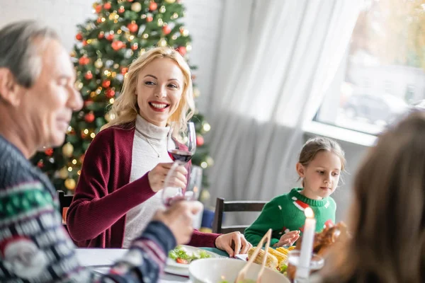 Allegra donna bionda brindare con un bicchiere di vino rosso vicino a padre e figlie offuscati a cena di Natale — Foto stock