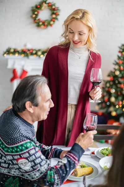 Heureuse femme blonde avec un verre de vin rouge grillé près père aîné pendant le dîner de Noël dans le salon — Photo de stock