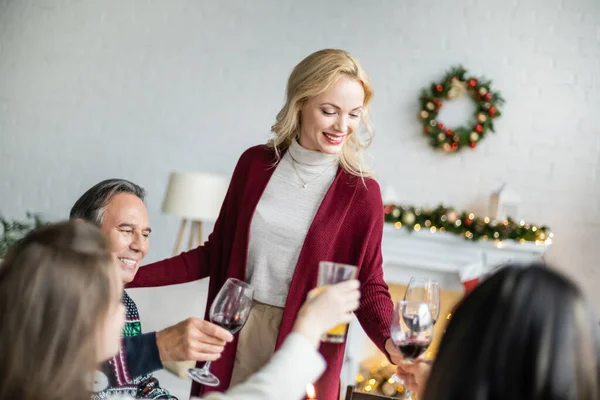 Mujer alegre de pie cerca de tostadas familiares con copas de vino mientras celebra la Navidad en casa - foto de stock