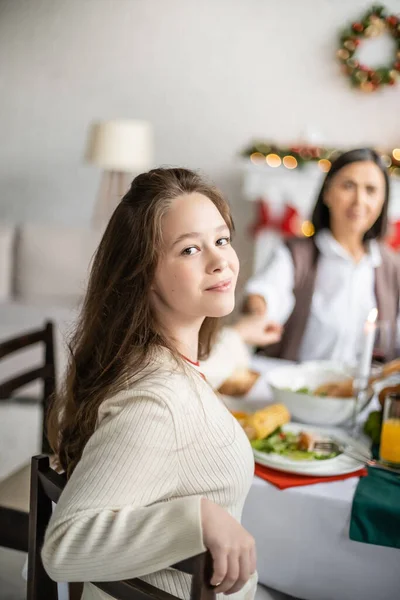 Preteen girl smiling at camera while holding hands with blurred multiracial granny during christmas dinner — Stock Photo