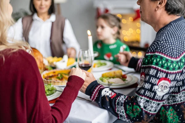 Família multiétnica de mãos dadas e rezando perto do jantar festivo durante a celebração do Natal — Fotografia de Stock