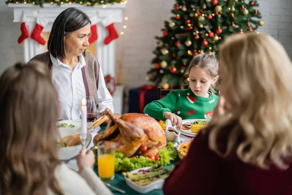 Mujer multirracial mirando a la nieta durante la cena festiva con la familia borrosa cerca del árbol de Navidad decorado - foto de stock