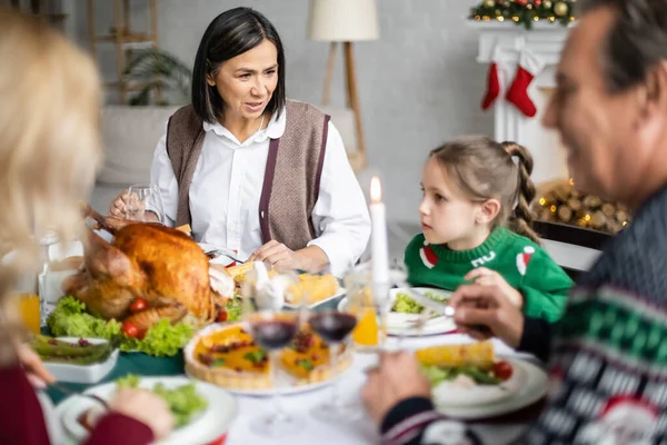 Multiracial granny talking to granddaughter during christmas dinner with blurred family — Stock Photo