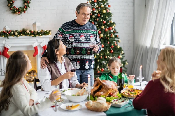 Vieillard griller avec un verre de vin rouge près de la famille multiethnique pendant le dîner de Noël festif — Photo de stock