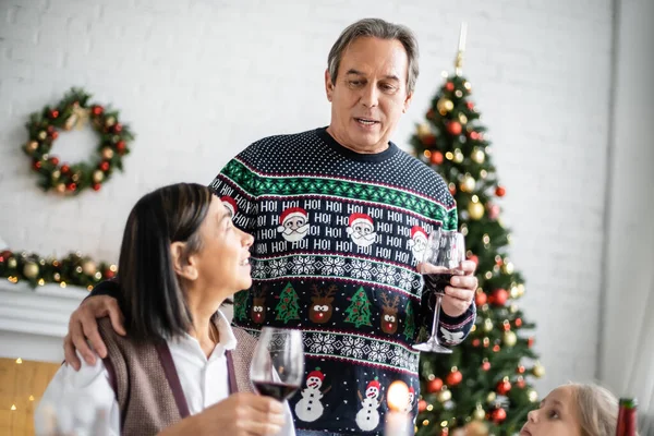 Hombre mayor abrazando a la esposa multirracial mientras brindar con un vaso de vino tinto cerca de su nieta durante la cena de Navidad - foto de stock