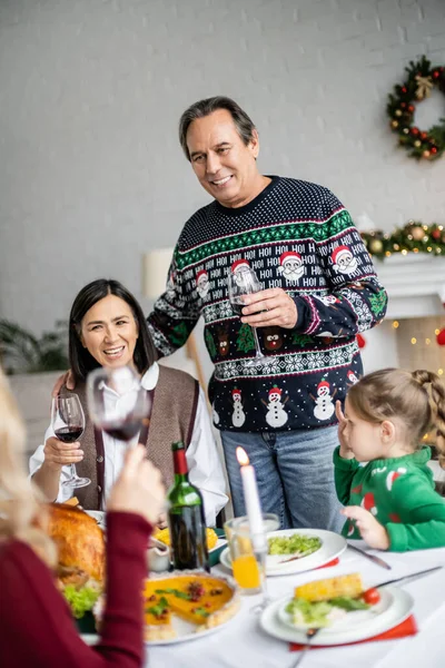 Heureux homme âgé griller avec un verre de vin rouge tout en célébrant Noël avec la famille interracial — Photo de stock