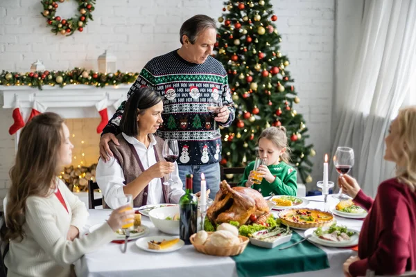 Hombre maduro tostando con copa de vino cerca de la familia interracial durante la cena de Navidad festiva - foto de stock