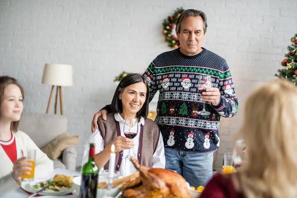 Homme âgé et femme multiraciale griller avec des verres de vin rouge sur le dîner de Noël avec la famille floue — Photo de stock