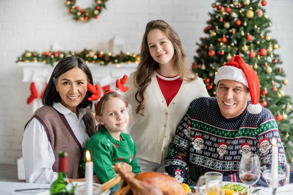 Fröhliche Mädchen mit multiethnischen Großeltern, die in die Kamera schauen, in der Nähe von verschwommenem Festmahl und geschmücktem Weihnachtsbaum — Stockfoto