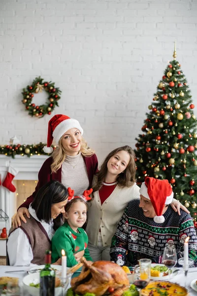 Glückliche Frau mit Weihnachtsmütze blickt in die Kamera in der Nähe multiethnischer Familien und geschmückter Weihnachtsbäume — Stockfoto