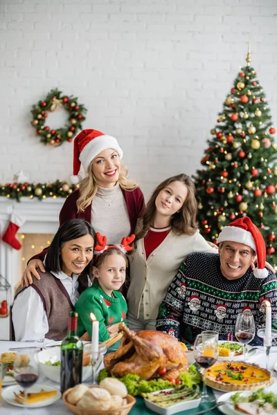 Familia multiétnica feliz mirando a la cámara cerca de la cena festiva y el árbol de Navidad en casa - foto de stock
