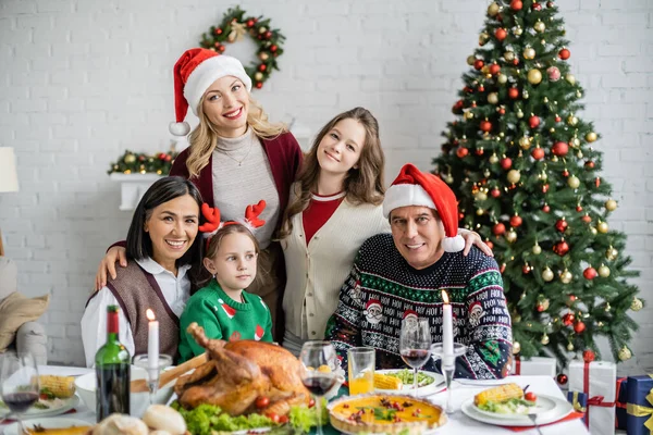 Familia multiétnica feliz mirando a la cámara cerca de deliciosa cena y árbol de Navidad - foto de stock