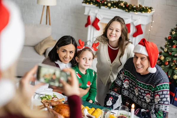 Mujer borrosa tomando fotos de hijas con abuelos interracial cerca de la cena festiva — Stock Photo