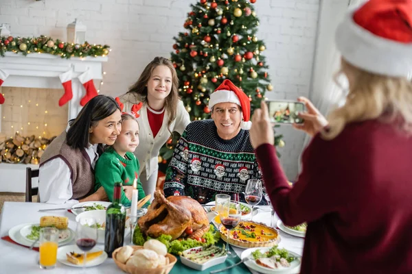 Mujer borrosa tomando fotos de la familia interracial feliz cerca de la cena festiva y el árbol de Navidad - foto de stock
