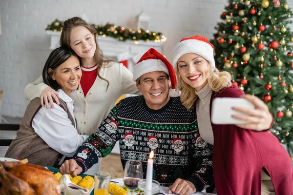 Mujer sonriente en sombrero de santa tomar selfie con familia multiétnica cerca de la cena festiva y árbol de Navidad - foto de stock