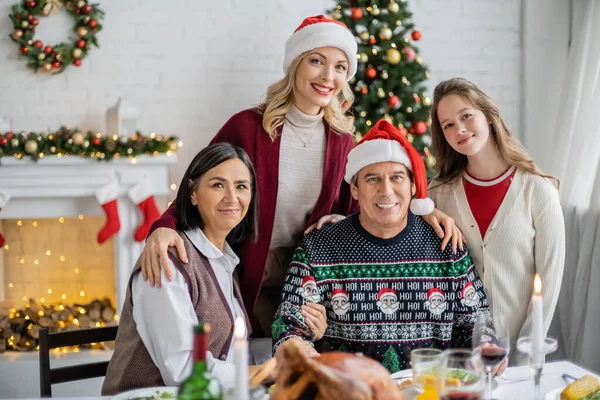 Glückliche Frau mit Weihnachtsmütze, die in der Nähe von Familie und Weihnachtsbaum in die Kamera schaut — Stockfoto