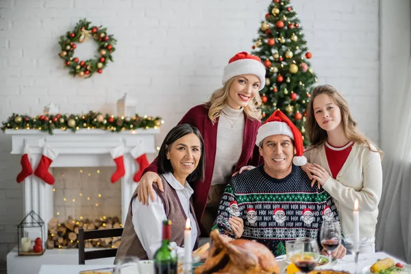 Feliz familia interracial sonriendo cerca de la cena festiva en la sala de estar con chimenea decorada y árbol de Navidad - foto de stock