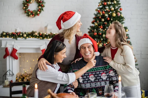 Familia multiétnica feliz riendo cerca de la cena festiva y decorado árbol de Navidad en la sala de estar - foto de stock