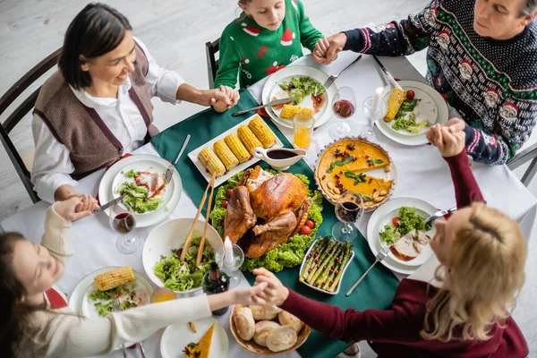 Blick von oben auf die multiethnische Familie, die sich die Hände hält und vor dem Erntedankdinner in der Nähe des köstlichen Essens betet — Stockfoto