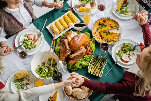 Vista dall'alto della famiglia interrazziale che si tiene per mano mentre prega vicino a gustoso pasto servito per la celebrazione del Ringraziamento — Foto stock