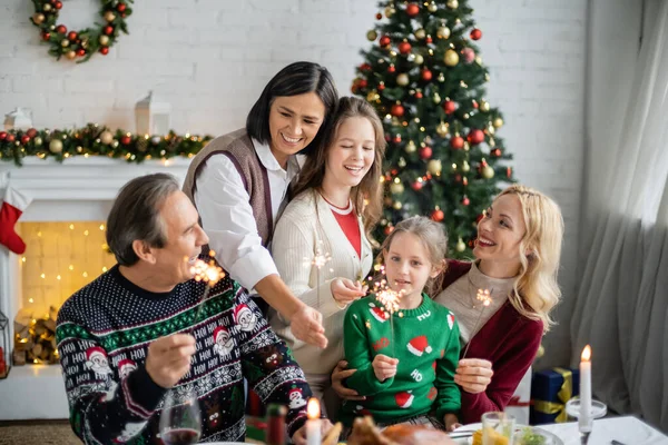 Familia multiétnica emocionada celebración de bengalas en la sala de estar con chimenea y árbol de Navidad decorado - foto de stock