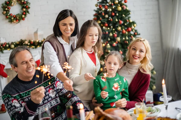 Alegre família multicultural segurando sparklers perto de jantar festivo na sala de estar com árvore de natal decorada — Fotografia de Stock