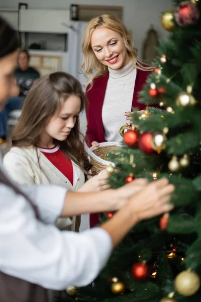 Blonde woman smiling near teen daughter and blurred multiracial mother decorating christmas tree — Stock Photo