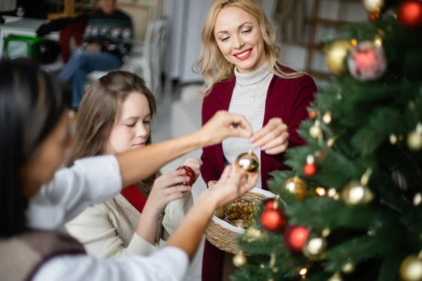 Mujer feliz decorando árbol de navidad con hija y madre multirracial borrosa - foto de stock