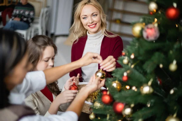 Fröhliche Frau mit Blick auf verschwommene multiethnische Mutter, die zu Hause den Weihnachtsbaum schmückt — Stockfoto